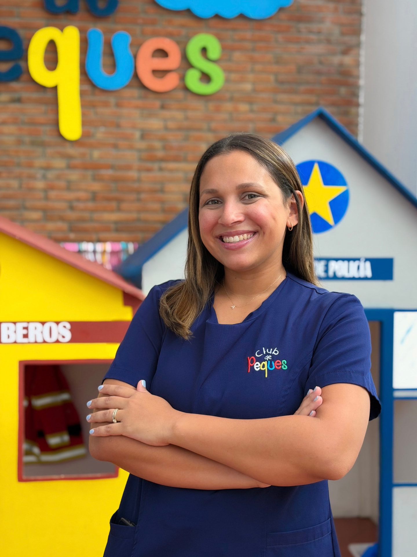 Portrait of a smiling woman with medium-length brown hair, wearing a blue scrub top with a logo. She is standing with arms crossed in front of a colorful indoor play structure.