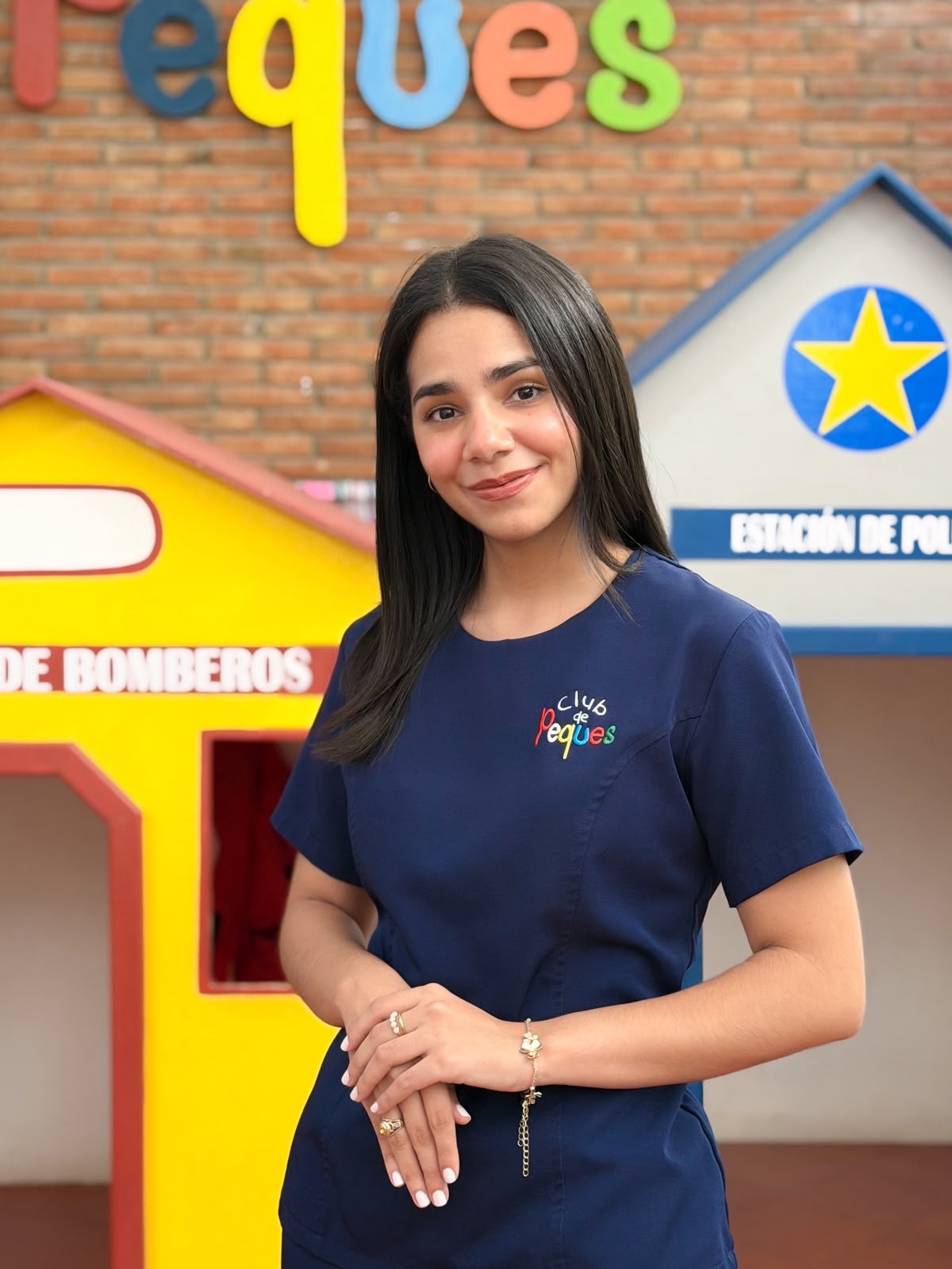 Portrait of a smiling young woman with long dark hair, wearing a blue scrub top with a logo. She is standing with her hands lightly clasped in front of a colorful indoor play structure.