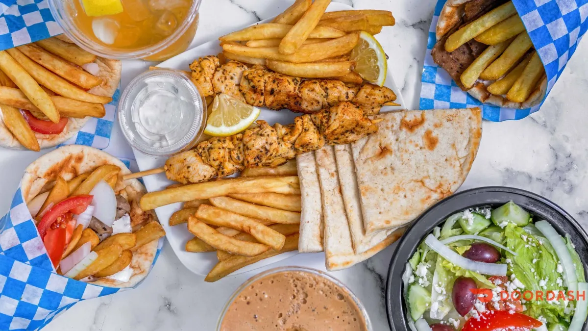 Overhead view of a Greek fast-food meal from Souvlaki Ville featuring chicken souvlaki skewers with lemon wedges, golden fries, warm pita bread, tzatziki sauce, Greek salad with feta and olives, and assorted gyro wraps with fries and fresh vegetables, served on a marble surface with iced drinks.