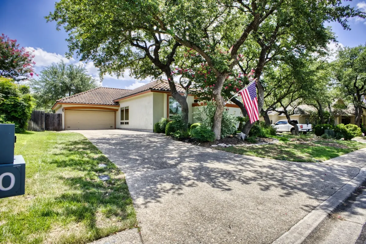 Front view of a suburban home with a wide concrete driveway, mature trees, a well-maintained lawn, and an American flag displayed near the entrance.