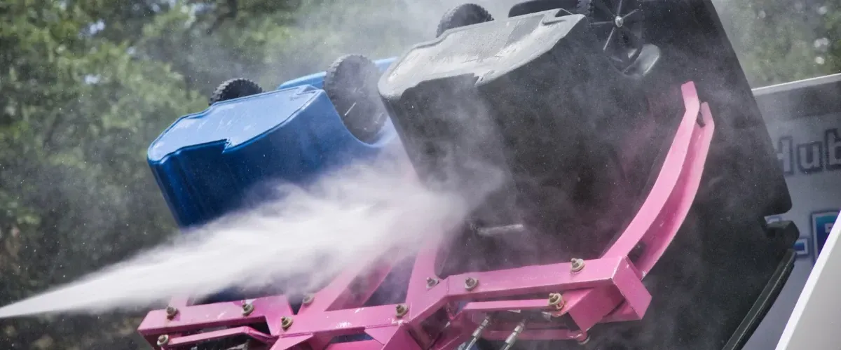 Trash bin cleaning High-pressure water jets cleaning trash and recycling bins using an automated bin washing systemlifting two bins for sanitizing, with a technician standing beside the equipment.