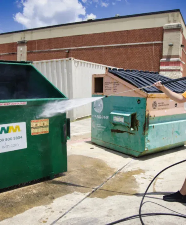 Commercial dumpster being cleaned with a high-pressure washer behind a building.