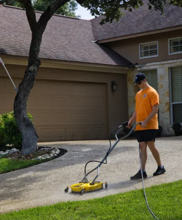Technician pressure washing a residential driveway in front of a suburban home.