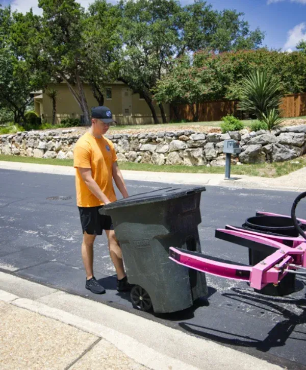A young worker in an orange shirt and black hat rolls a green trash bin towards a lifting mechanism on a sunny street.
