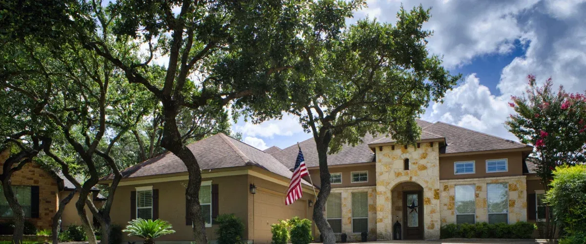 A large stone and stucco house with a brown roof and arched entryway, nestled among green trees under a cloudy sky.