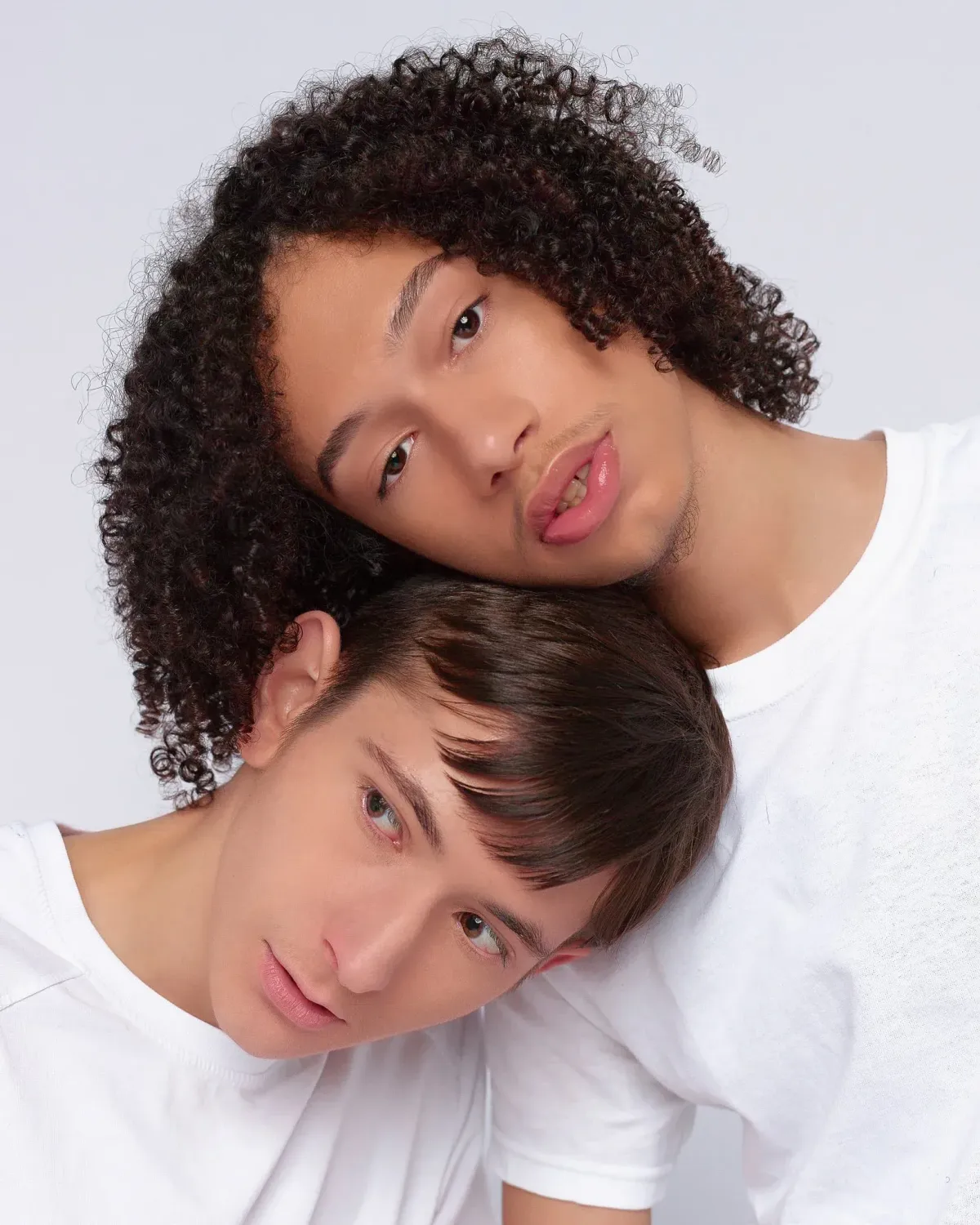 Studio portrait of two young men posing closely together. The top man is resting his chin near the head of the lower man. Both wear plain white t-shirts against a plain white studio backdrop. Warm, natural lighting.