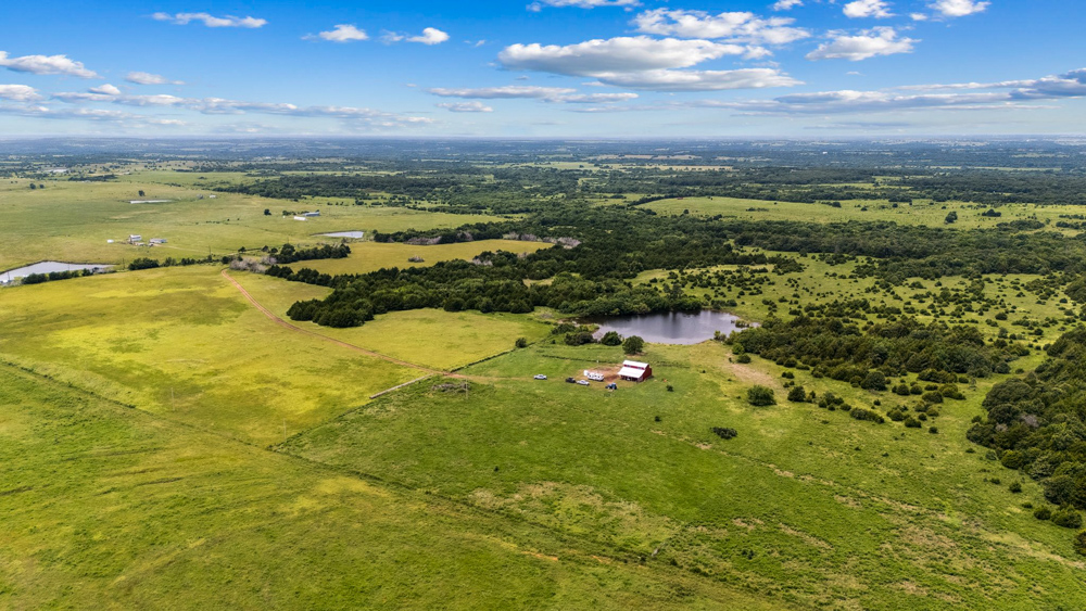 Aerial view of 108-acre Rock n Wood Farm & Ranch for sale in Pawnee, Oklahoma, showing open pastureland, pond, trees, and a red barn surrounded by rolling countryside.