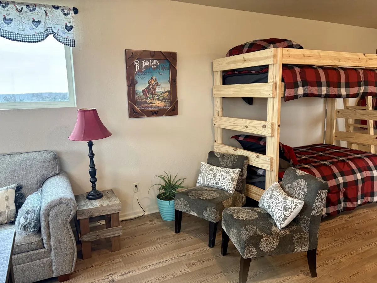 Rustic cabin bedroom with custom wooden bunk beds, plaid bedding, cozy seating area, side table with lamp, and farmhouse decor.