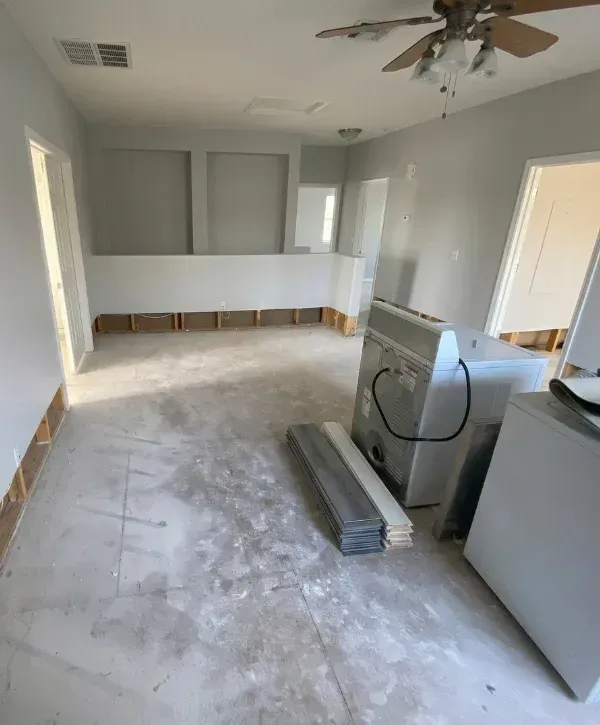 A wide shot of an empty room undergoing renovation, featuring exposed subfloor, white walls, and stacks of new hardwood flooring planks.