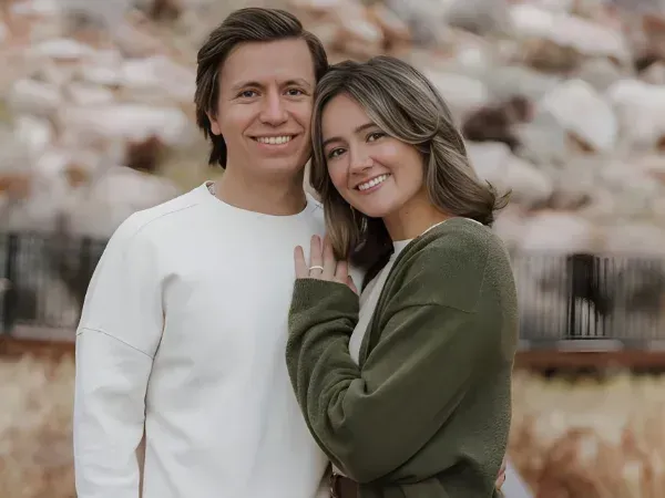 A smiling young couple poses happily outdoors against a blurred natural rock background.