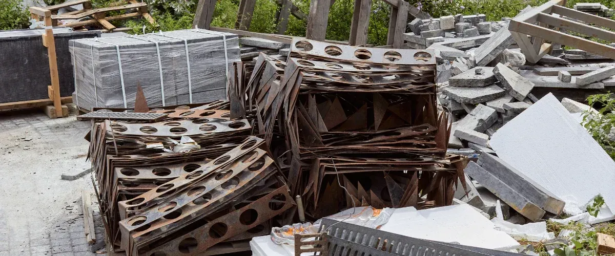 A cluttered view of construction waste, including rusty metal sheets, wrapped paving stones, and rubble on a dirt ground.