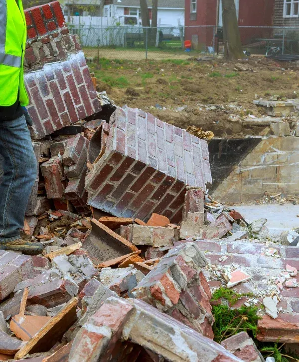 A construction worker in a safety vest stands beside a pile of fallen bricks and rubble at a demolition site.