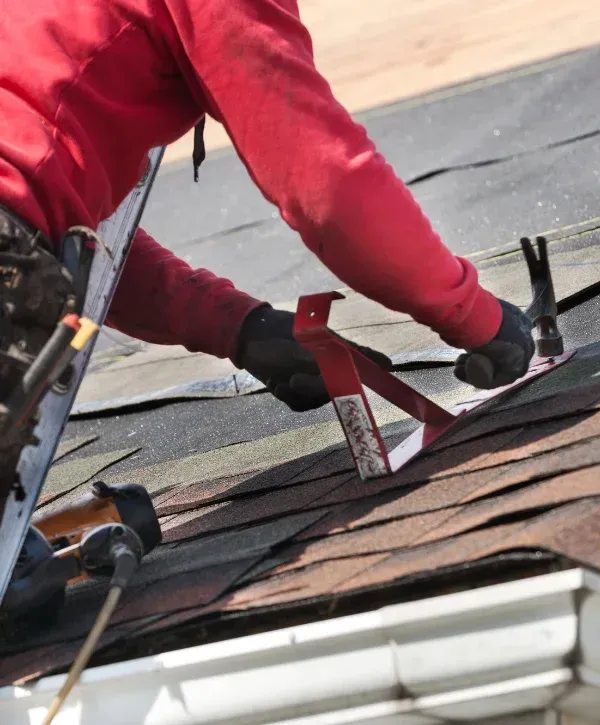 Close-up of a roofer in a red shirt and black gloves installing shingles on a roof with a hammer.