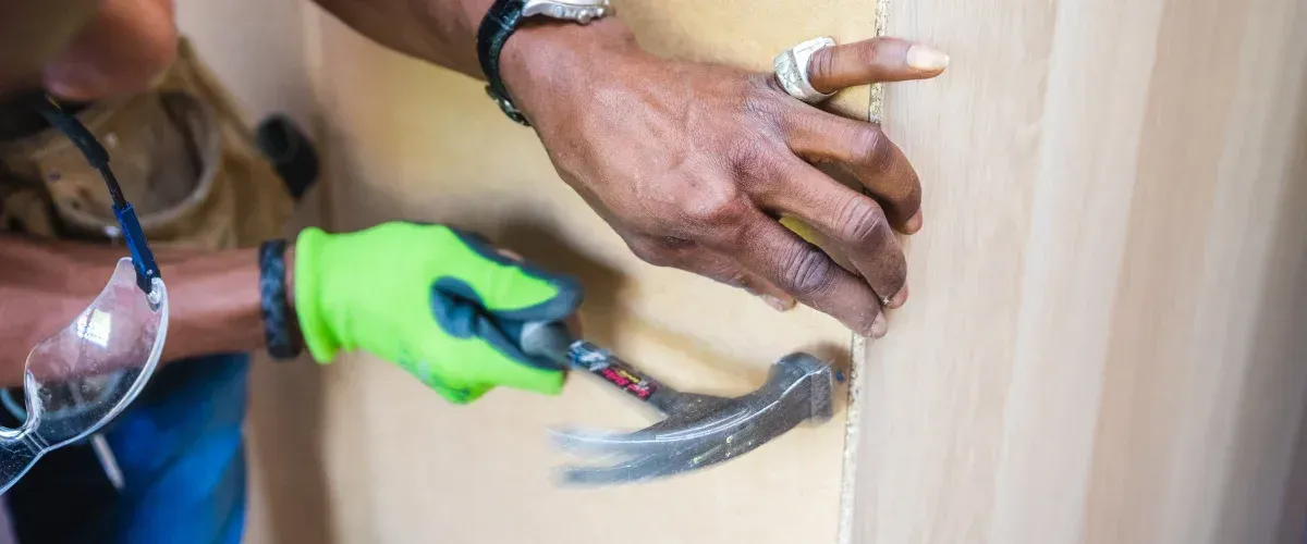 A craftsman in bright green gloves hammers a nail into light wood, with safety glasses hanging nearby.