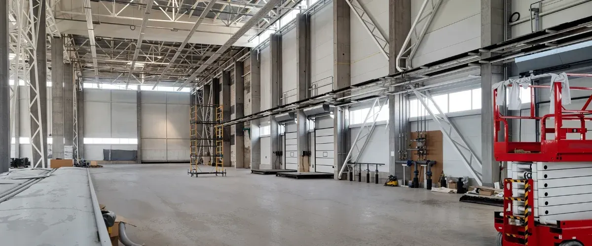 A wide, empty industrial warehouse interior with concrete pillars, white walls, and an overhead crane system under construction.