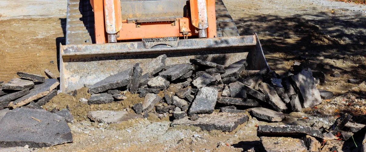 An orange excavator bucket filled with broken pieces of black asphalt on a dirt ground at a construction site.