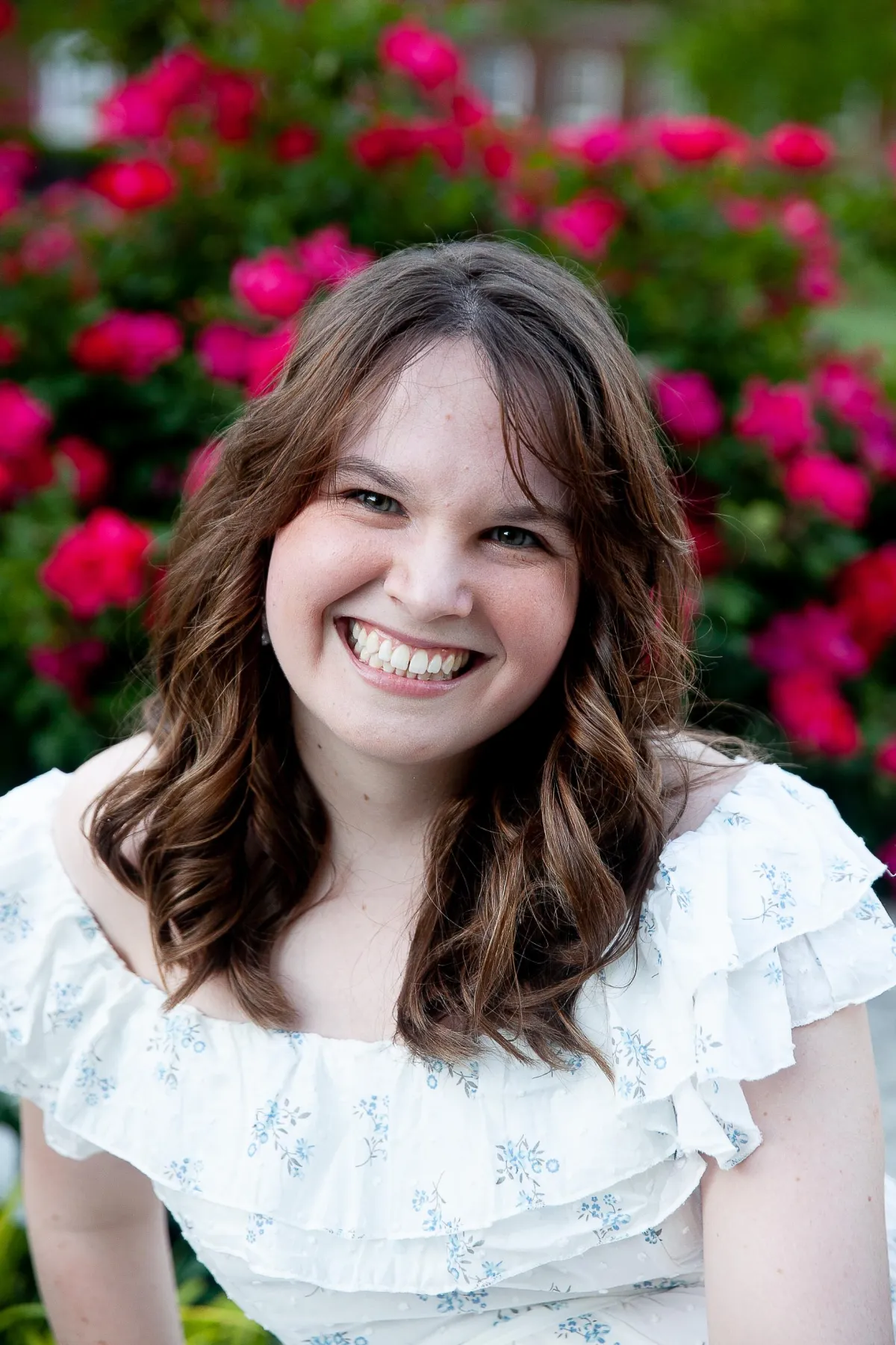 picture of the founder in a white dress with blue flowers, brown hair, green eyes smling in front of a wall of flowers