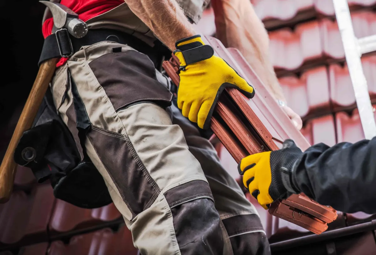 Two roofers wearing yellow gloves and tool belts carrying metal shingles onto a roof