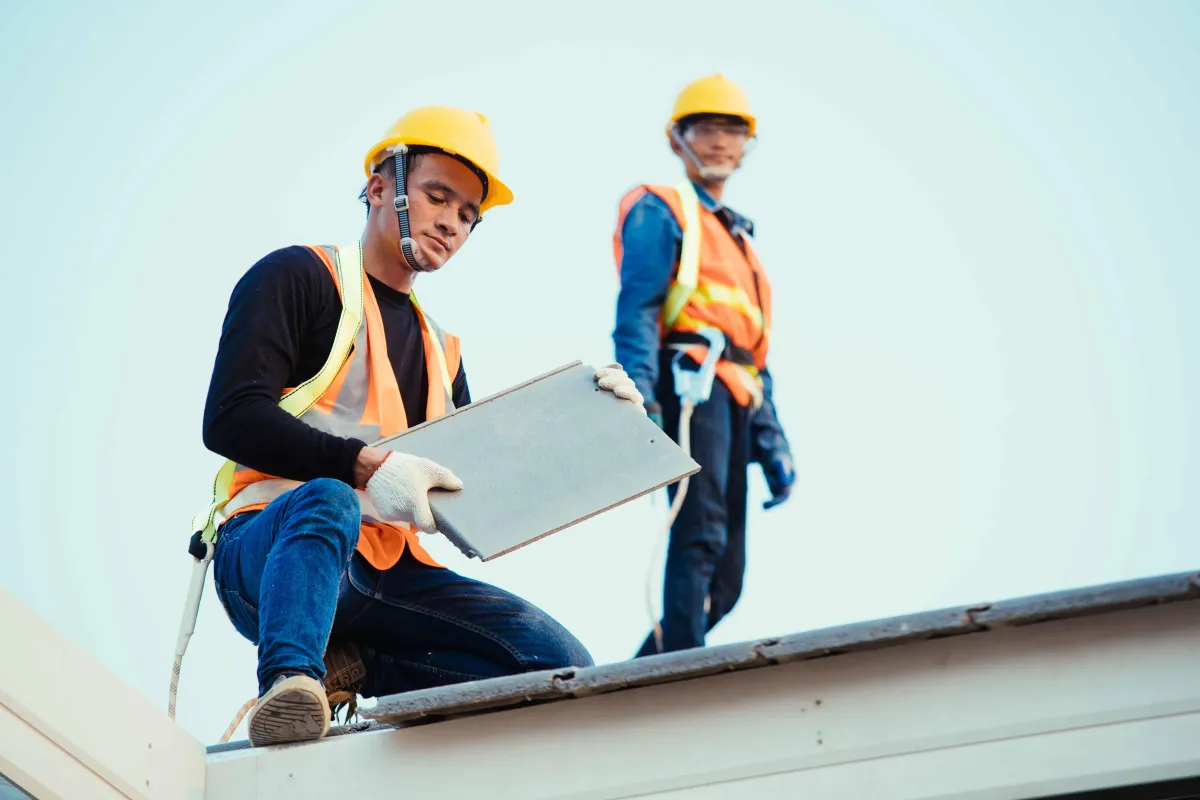 Two construction workers wearing yellow hard hats and orange safety vests standing on flat roof inspecting roof tile