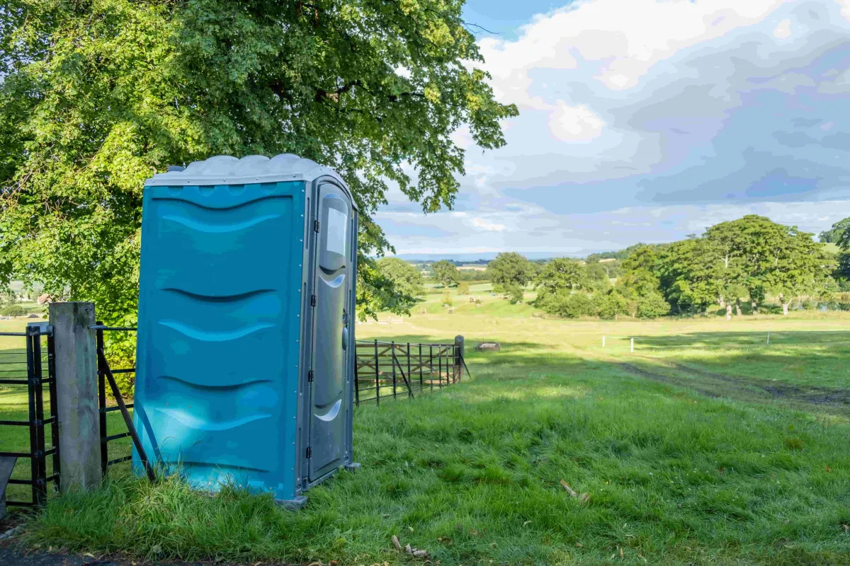 One blue porta potty in front of a small fence and big tree in a fielded area 