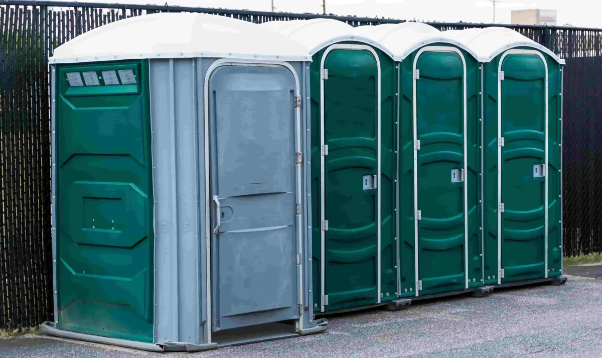 Four green porta potties lined up side by side on cement in front of a metal fence