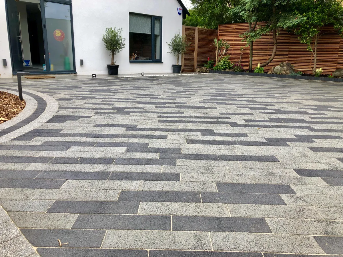 The driveway features an elegant design with interlocking pavers, providing a smooth and durable surface for vehicles. Lush greenery and colorful flowers border the driveway, enhancing its aesthetic appeal and blending harmoniously with the surrounding natural environment.