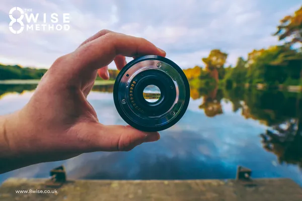 A person’s hand holding a camera lens up to a serene lake and forest landscape, symbolizing the 8Wise® Method lens and gaining clarity on life events and wellbeing.