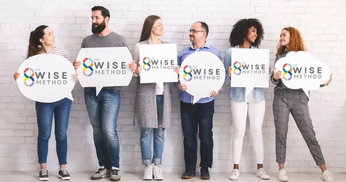 A diverse group of six people standing against a white brick wall, smiling and holding white speech bubbles and signs featuring 'The 8Wise® Method' logo. This visual represents the universal application and community aspect of the 8Wise® framework across different sectors of society
