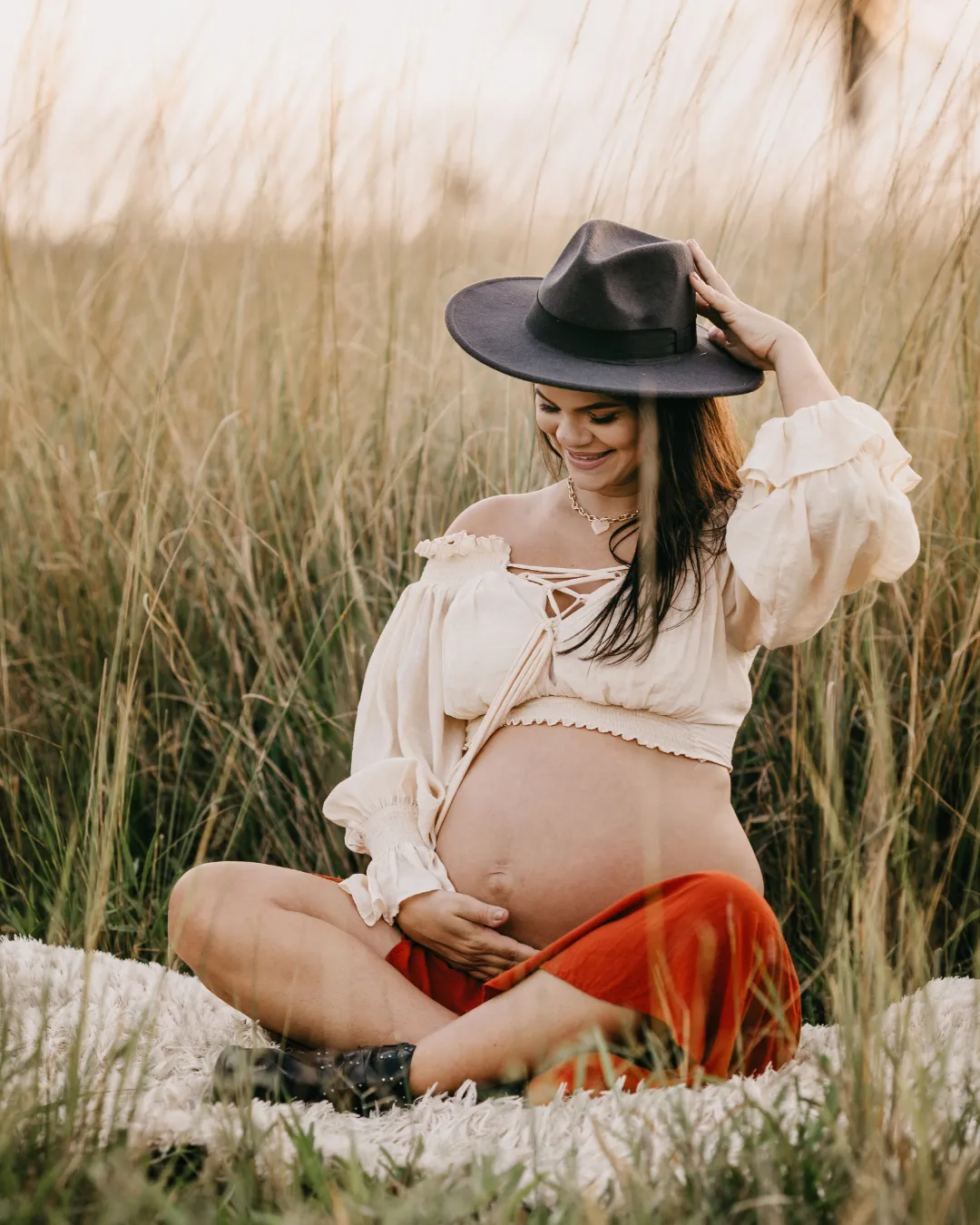 Pregnant woman sits cross legged within a grassy landscape, she is wearing a hat.She smiles and appears calm and confident