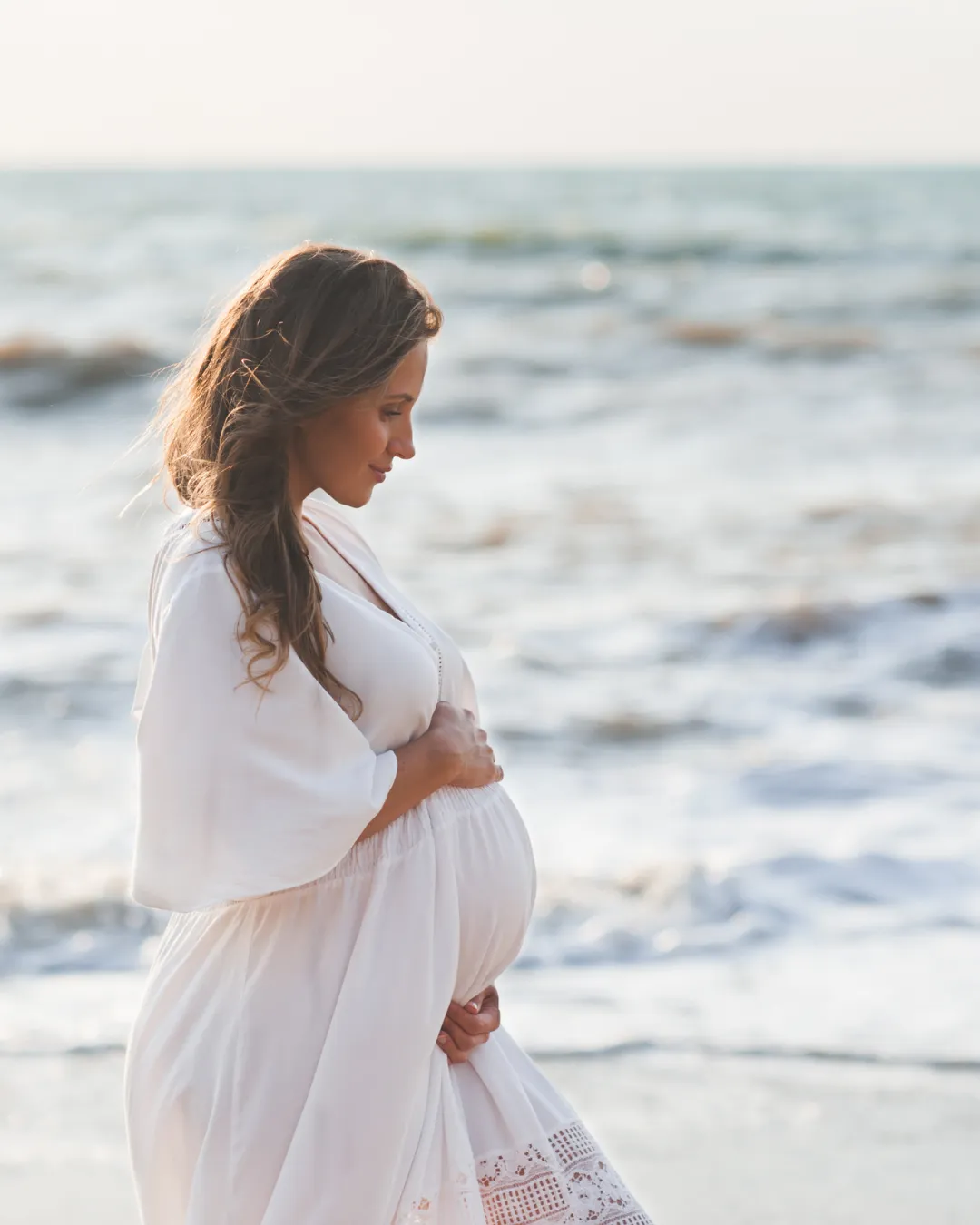 A pregnant woman walks along the shoreline, she wears white and her baby bump is visible. She smiles, and appears calm and confident