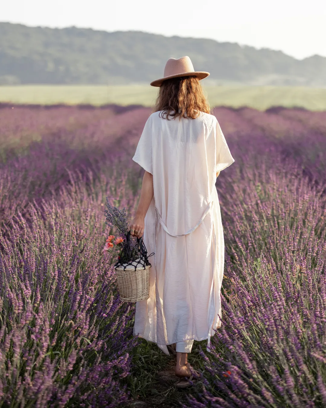 Pregnant woman walks through field o lavender, she carries a basket and is wearing a hat.She appears calm and confident