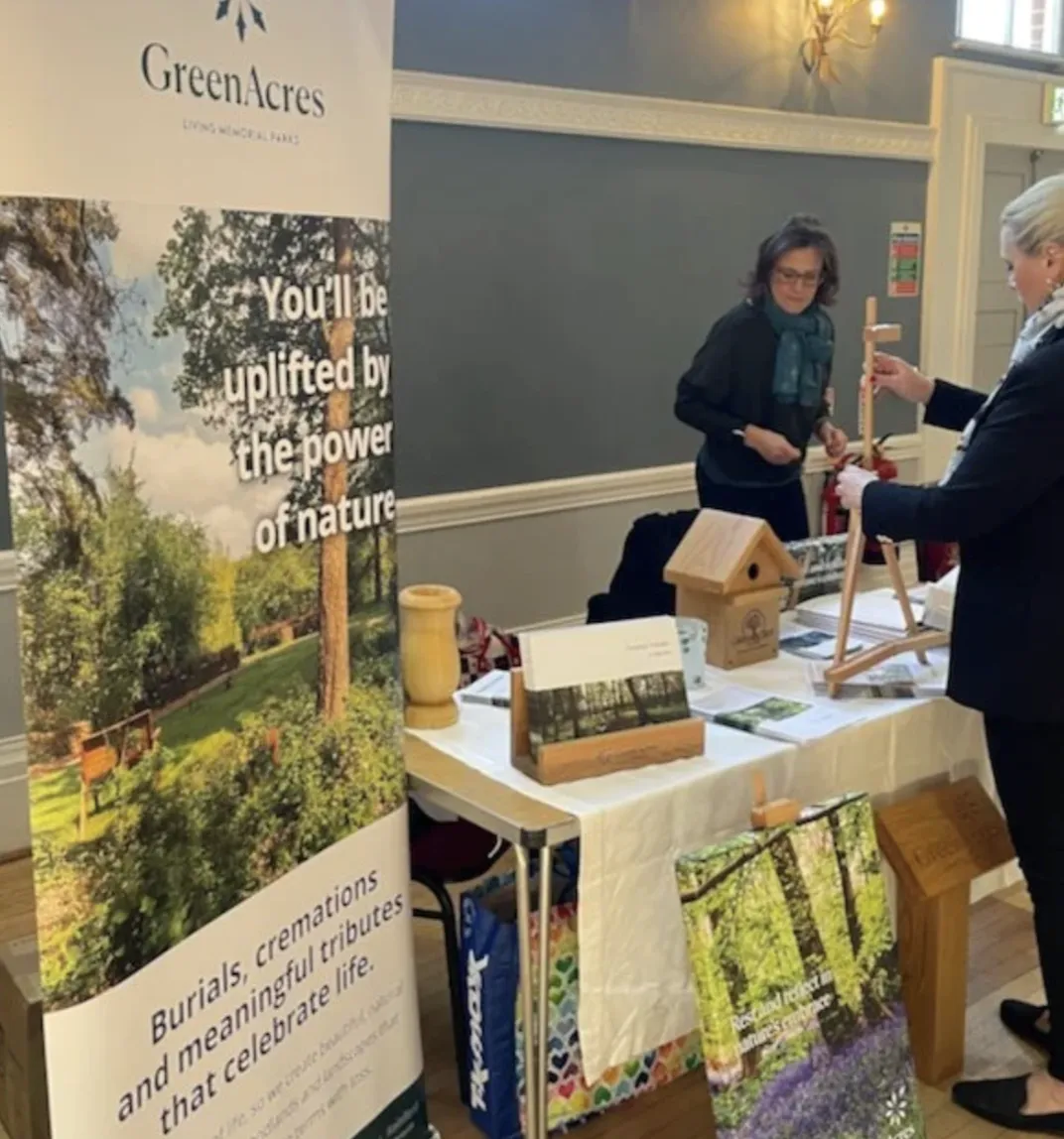 A booth at an indoor event, featuring a banner for Green Acres Living Memorials with a scenic outdoor photo. A woman stands behind the table, which displays wooden memorial items such as a birdhouse and a lantern. Another woman is interacting with the display, holding a wooden item. The table is covered with a white cloth and colourful bags are visible underneath.