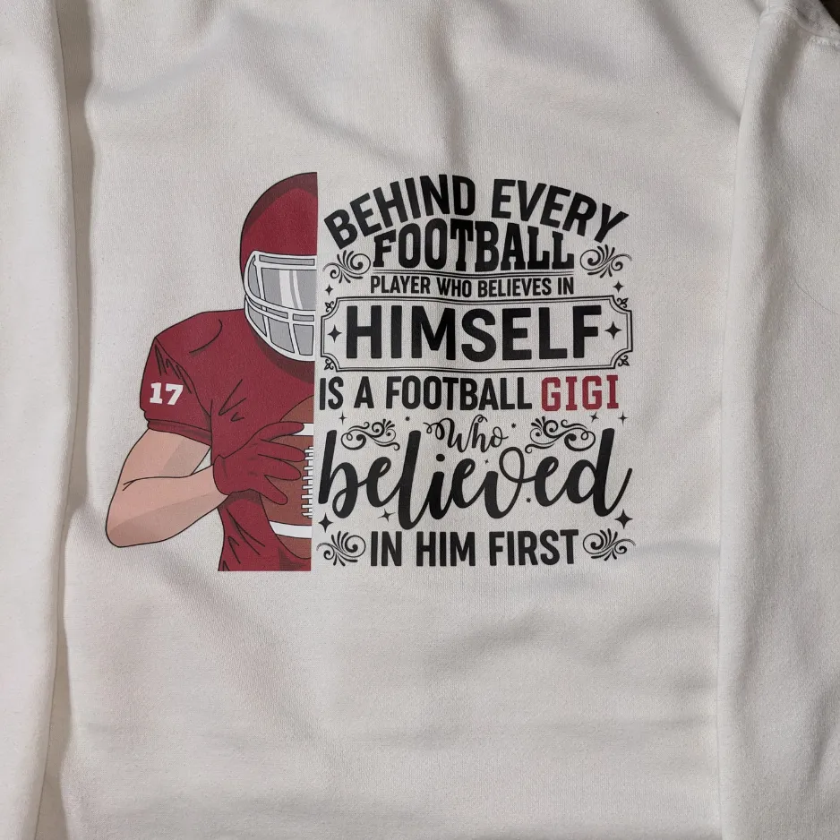 Custom printed white apparel featuring a maroon football player graphic and the text 'Behind Every Football Player Who Believes In Himself Is A Football Gigi Who Believed In Him First'