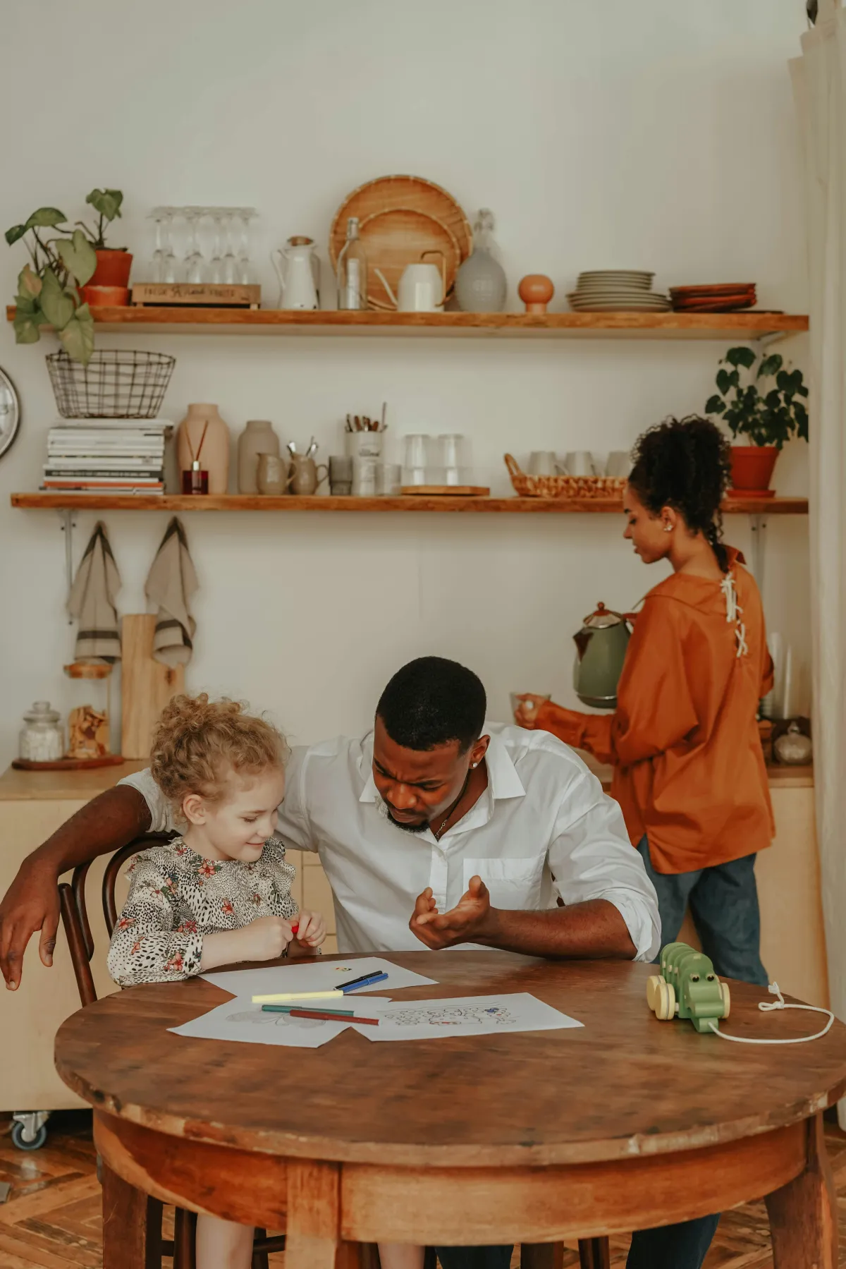 A family in the kitchen with the dad helping their daughter with homework while the wife is pouring a drink