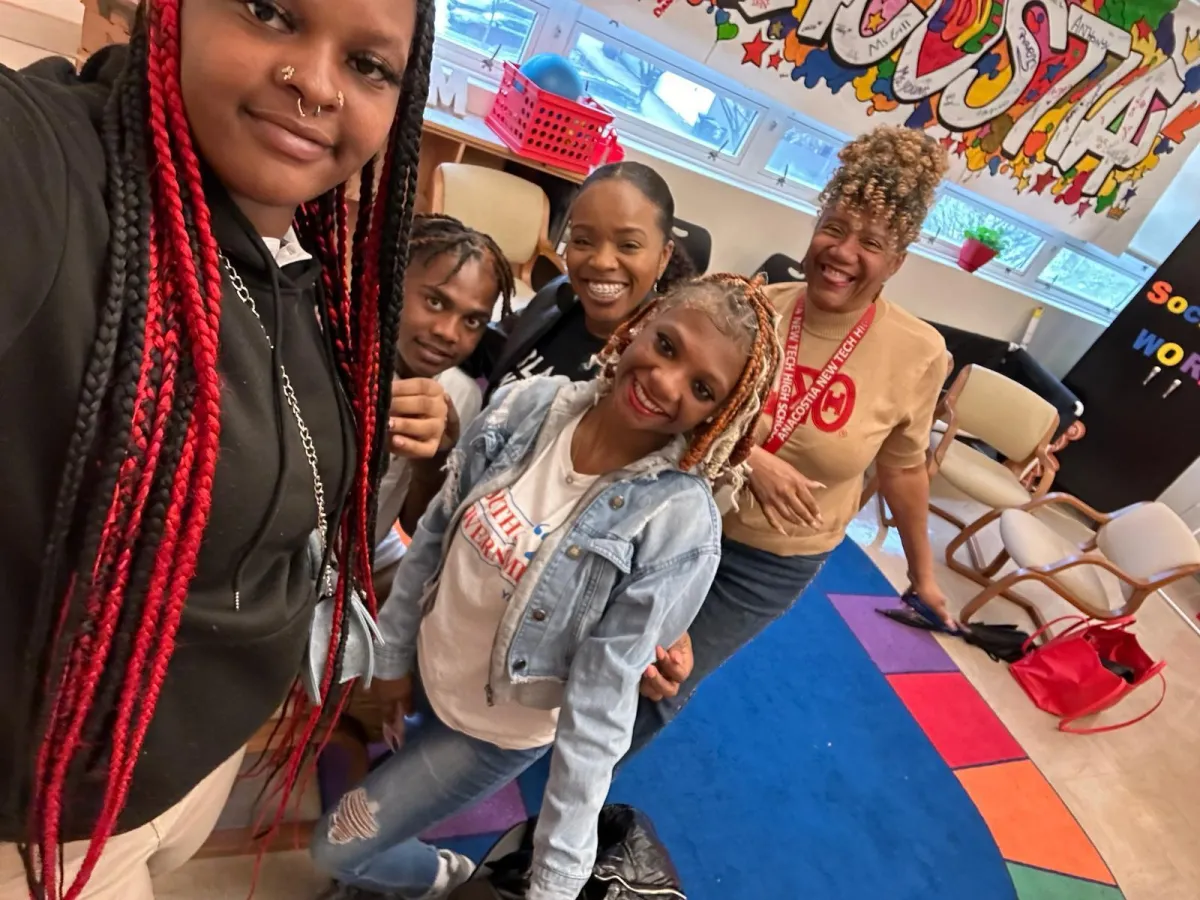 Koach Keisha posing with a group of young adults in a classroom setting, where she is speaking about the importance of self-confidence, time management, and making good choices. The group is smiling and standing closely together, creating a warm and engaging atmosphere. One young woman with red and black braids takes a selfie in the foreground, while others, including a teacher in a tan shirt and school lanyard, stand behind her. The classroom is decorated with colorful artwork, a vibrant rug, and chairs arranged for discussion, suggesting an interactive and supportive learning environment.