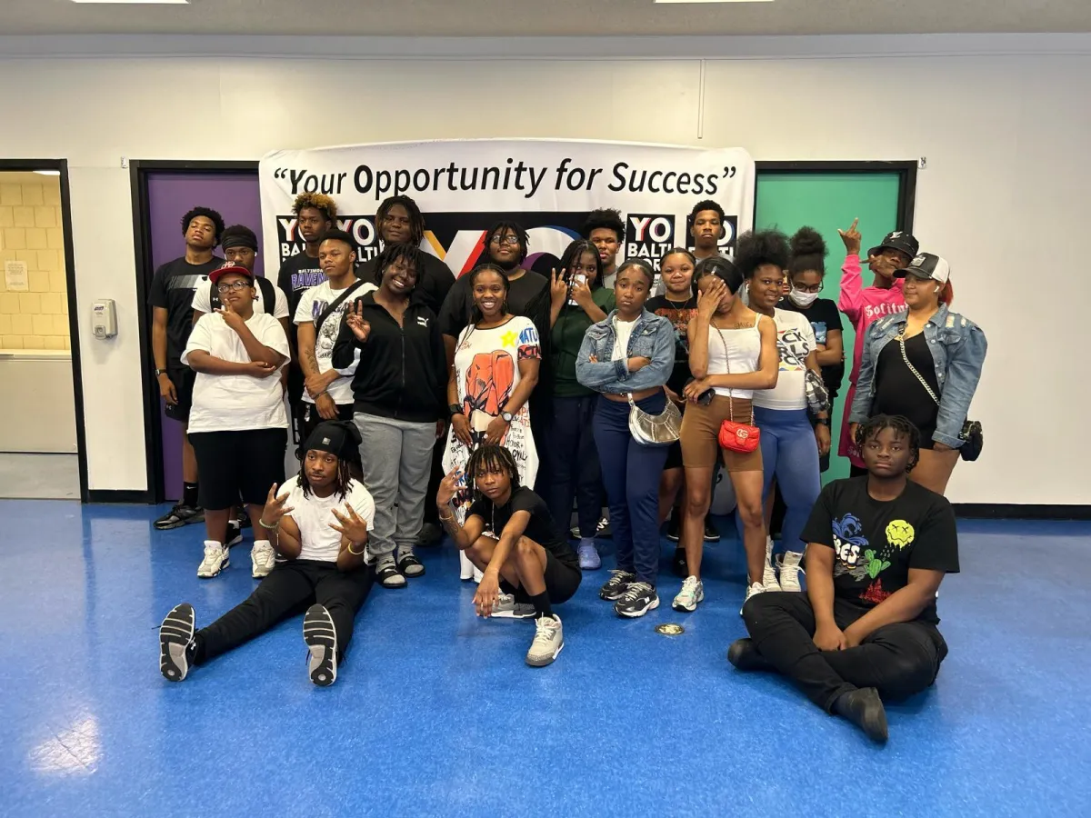 Koach Keisha posing with a diverse group of young adults at the 'Opportunity for Success' event at a local school. The group stands in front of a banner with the event's title, dressed in a mix of casual and sporty attire. Some participants are smiling, making hand gestures, or standing with arms crossed, showcasing confidence and camaraderie. The setting appears to be an indoor school or community space with a bright blue floor and colorful doors in the background.