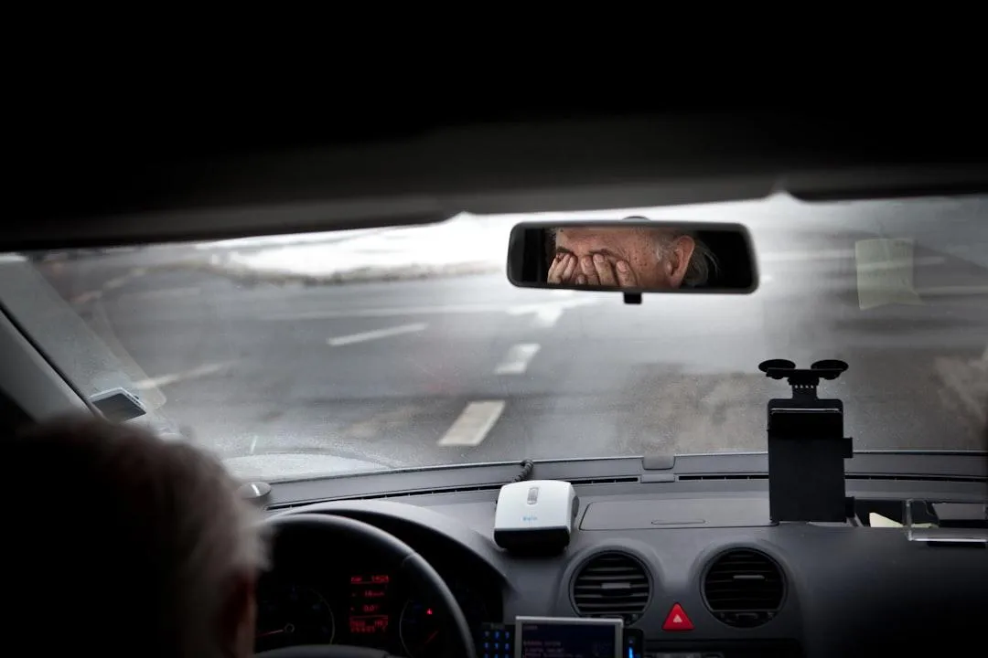 An older man sitting in the driver's seat of a car, rubbing his eyes while looking tired, seen through the rearview mirror.