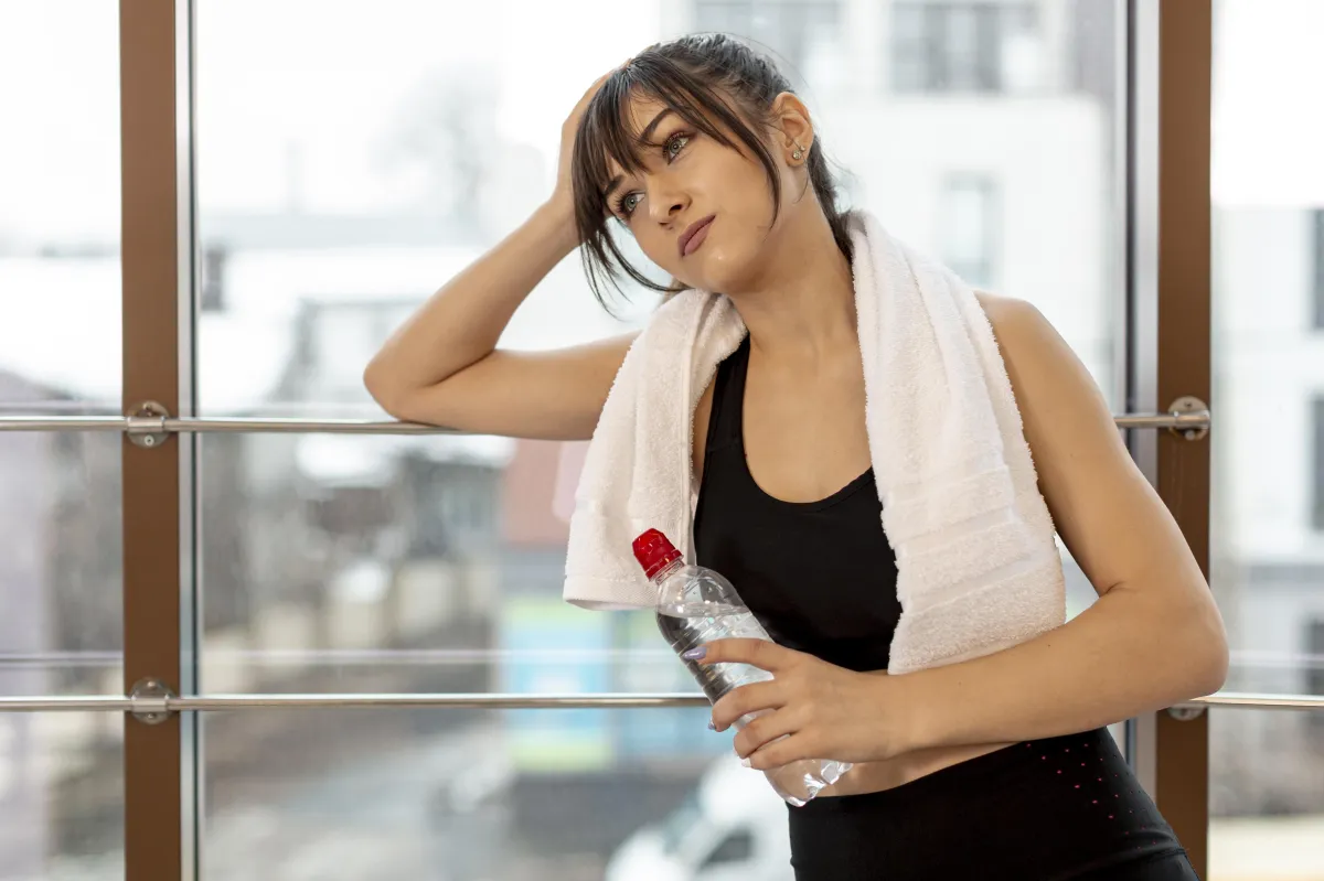 Tired woman with towel around neck holding water bottle.