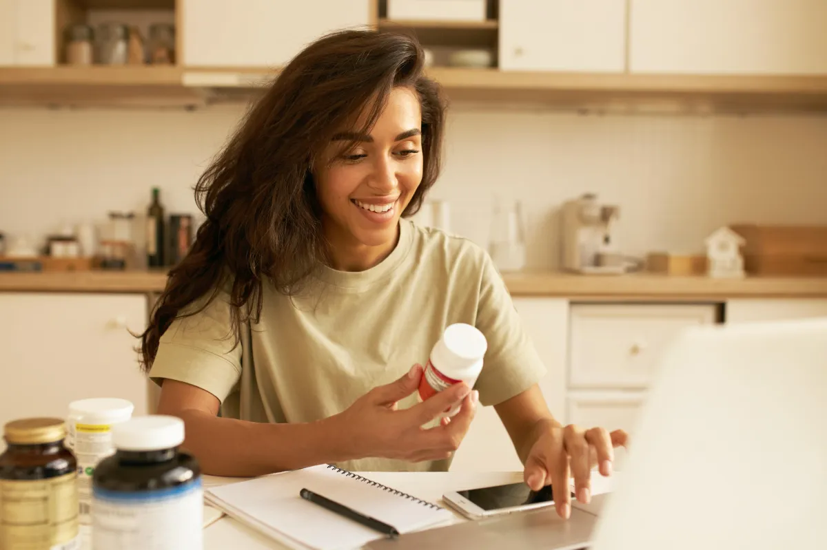 A woman sitting at a kitchen table, smiling and holding a medicine bottle, with several supplement bottles and a notebook on the table.