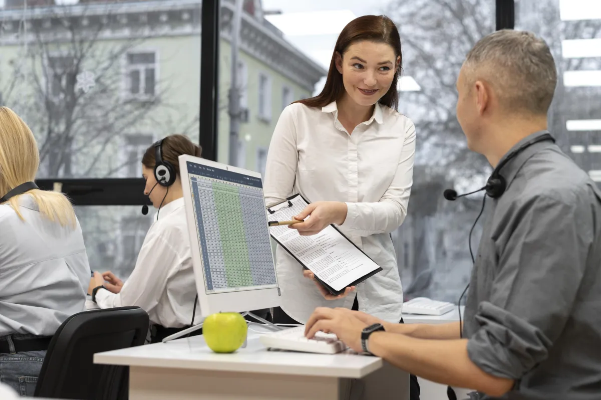 A woman standing and smiling while talking to a man seated at a desk with a computer monitor in an office setting, with other people working in the background.