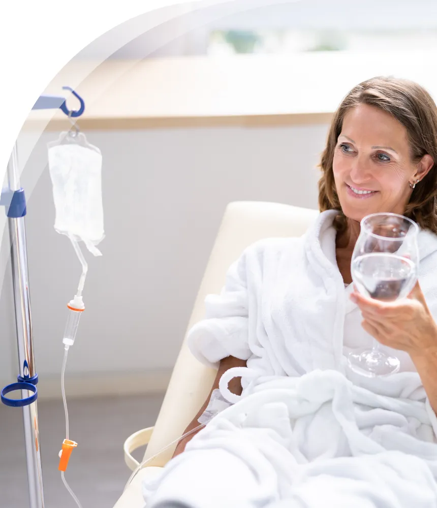 Woman receiving IV therapy at home, sitting comfortably in a robe and holding a glass of water.