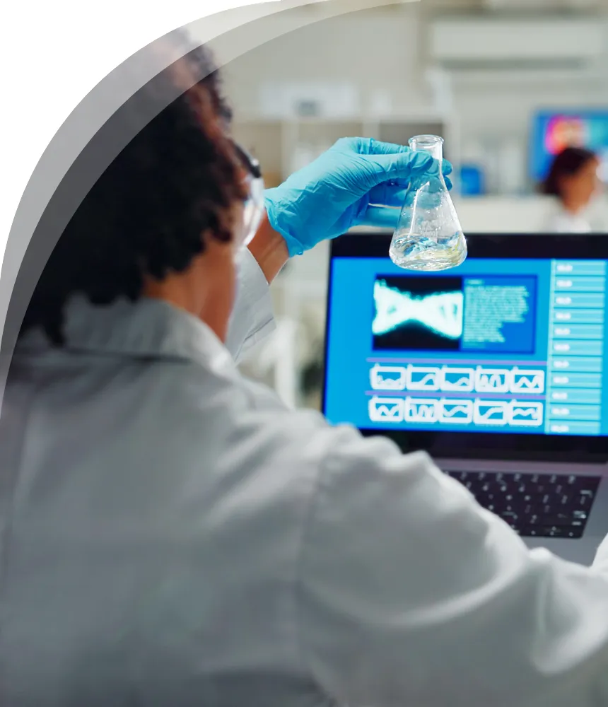 A scientist or researcher wearing blue gloves holding a flask with liquid, working in a lab with a laptop displaying scientific data in the background.