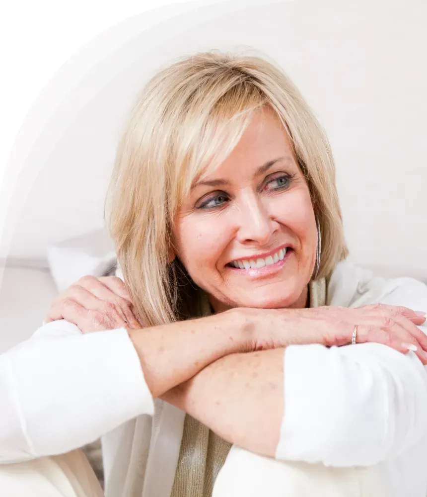 Smiling middle-aged woman resting her arms on a white surface, looking to the side thoughtfully.