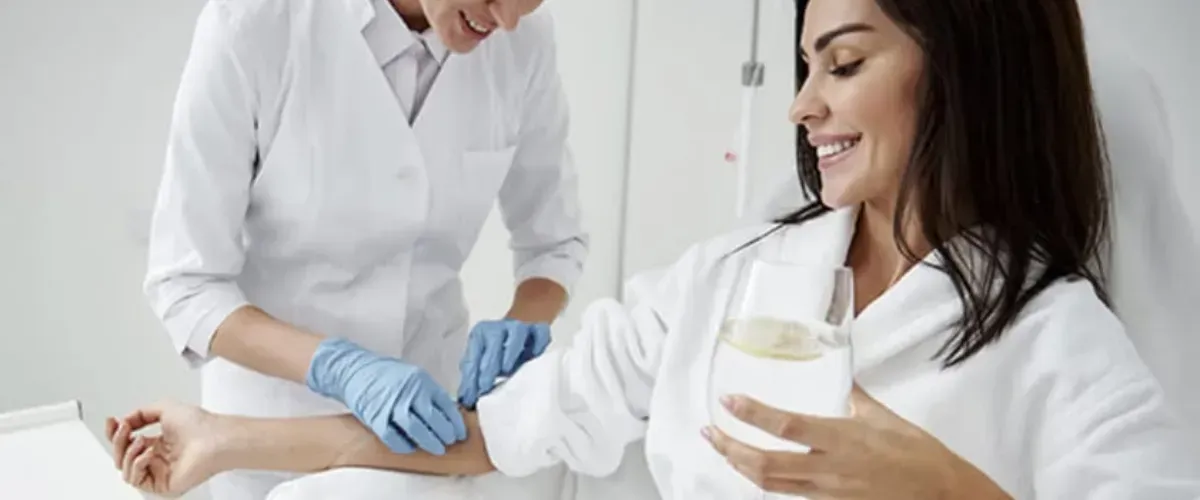 Smiling woman receiving IV vitamin therapy from healthcare professional while holding a glass of water