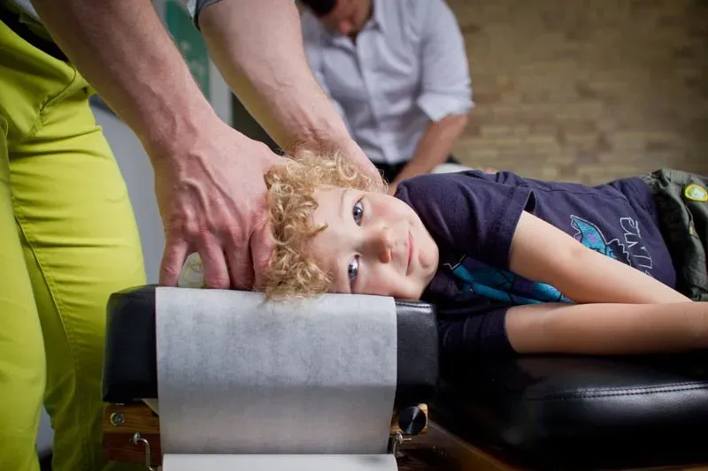 A young child with curly blonde hair lies on a chiropractic adjustment table, smiling while a practitioner gently holds their head.