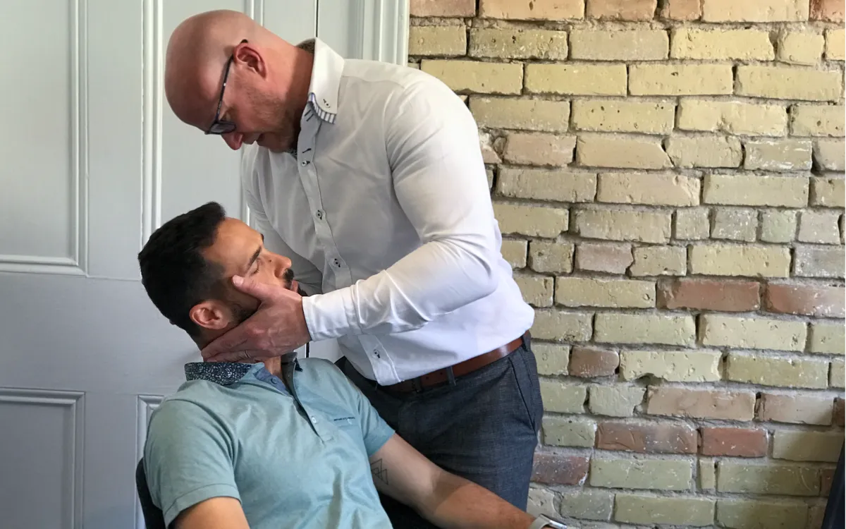 Dr. Richards performs a manual neck adjustment on a seated male patient in a clinical setting with a brick wall background.