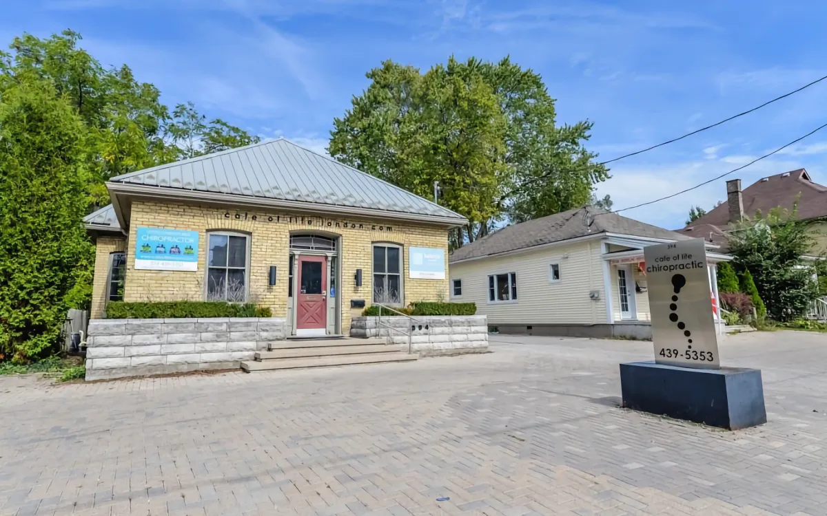 The exterior of Cafe of Life Chiropractic Studio shows a charming brick building with a red door, signage, and a modern signpost in the front parking area.