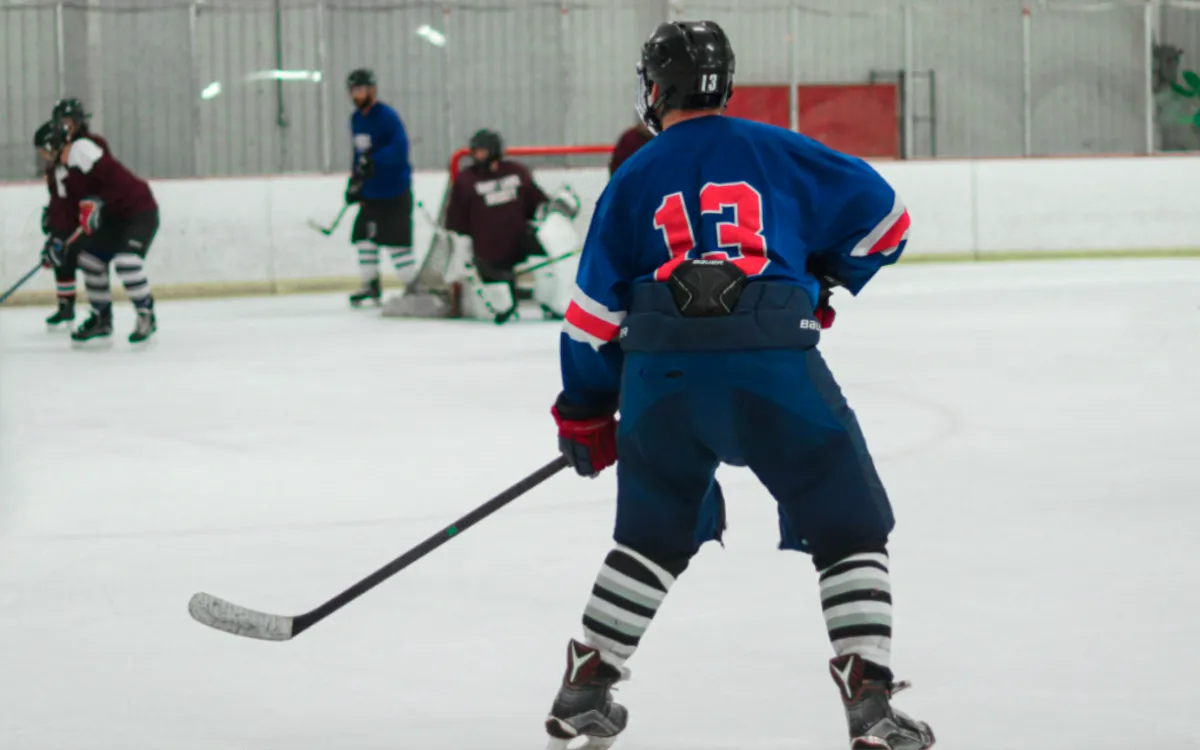 A hockey player wearing a blue jersey with the number 13 skates on the ice toward the goal as teammates and opponents gather near the net.