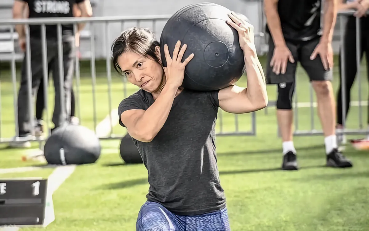 A woman in athletic clothing carries a heavy medicine ball on her shoulder during an outdoor fitness competition.