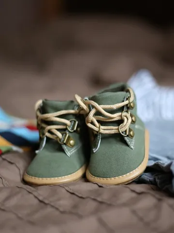 A pair of small green baby boots with tan laces sits on a soft brown blanket, surrounded by folded clothes.
